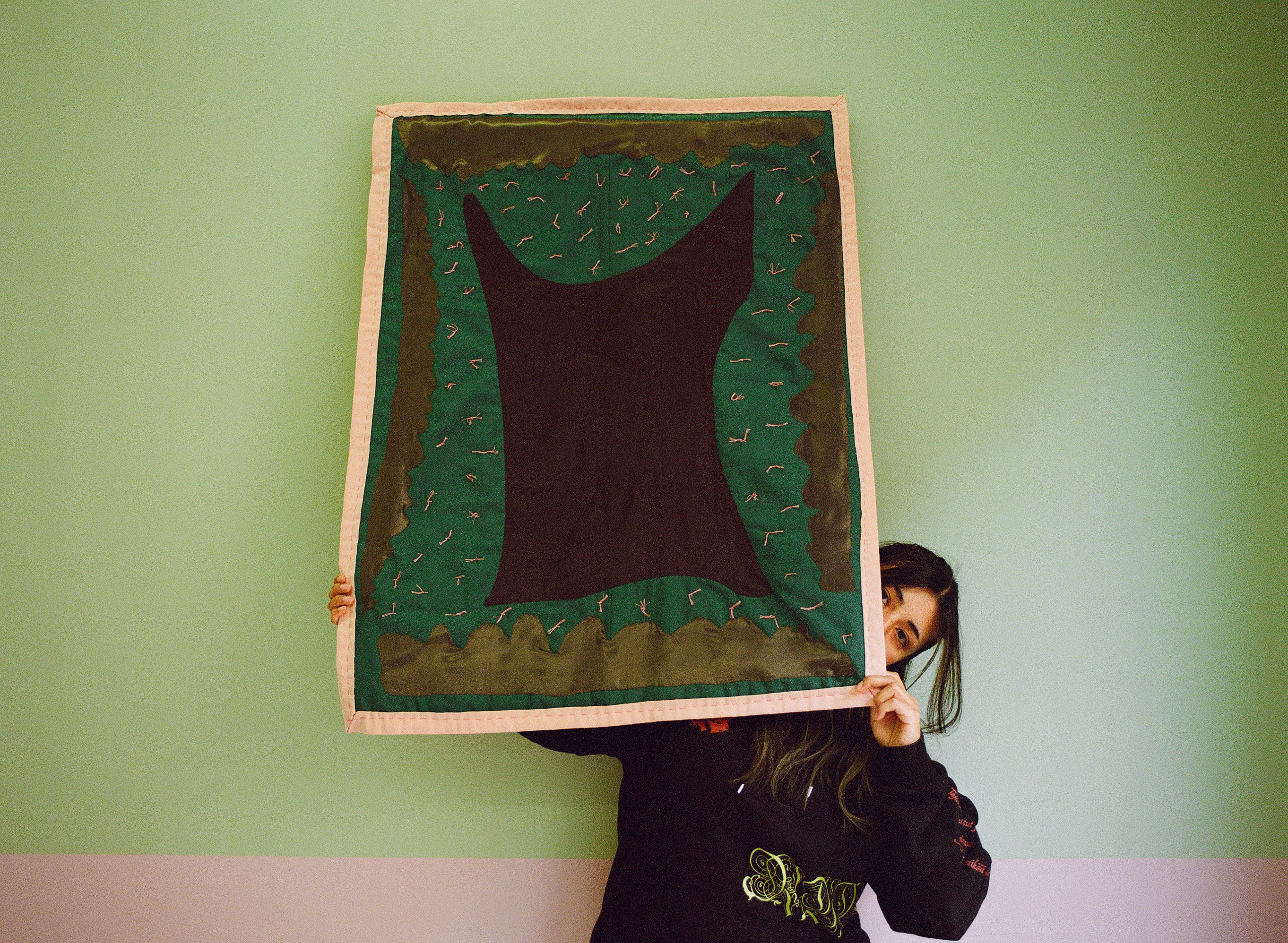 [Image description: A large quilt with organic shapes and hand-tied knots scattered throughout its soft surface hangs on the wall. The wall is a pale green with a dusty pink border running across it. The quilt has a light pink border with an emerald green background. The shapes within the background are dark purple and a mossy green. The hand-tied knots on the quilt are light pink. The artist stands behind the quilt, holding it up from its corners and peeking out from the left corner. Only her eyes are visible; she wears a black hoodie and long black hair.]
