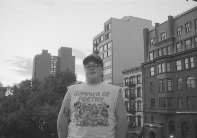 A black and white photograph of a white, fat person standing in front of some New York City buildings and trees. The image has been taken on an analogue camera and is grainy. The person wears glasses, a cap, and a white t-shirt with the sleeves cut off. The t-shirt reads ‘summer of poetry’ and features a selection of classic cartoon characters.
