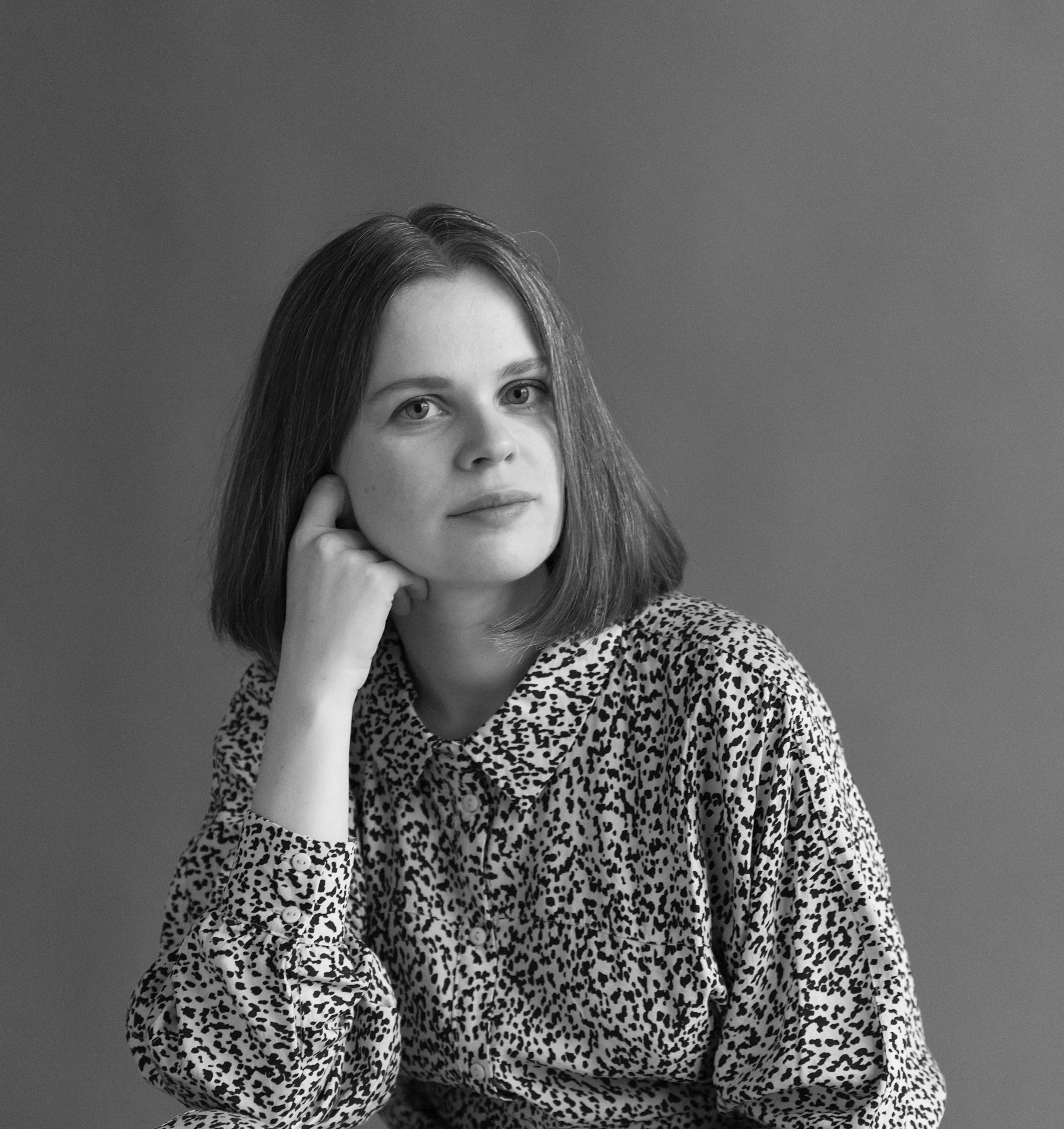 A black and white photograph of a person with shoulder-length hair wearing a printed button-down shirt, resting her right cheek on her hand