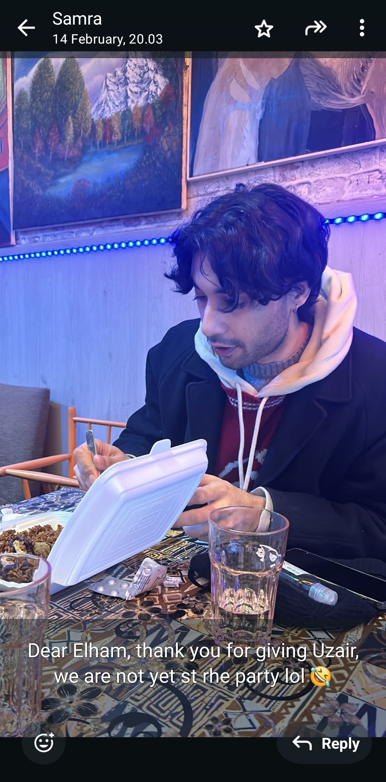 Image description: A screenshot of a message exchange between the artist's friends, Samra and Elham, dated 14 February 2025 at 20:03. The artist sits at a patterned table in a cozy café, eating from a Styrofoam takeaway box. He wears a black coat over layered winter clothing, with wavy hair falling around his face and a small earring in his left ear. The table holds two clear glasses, a beanie, antihistamines, and a hand sanitizer tube. Behind him, framed paintings of a snowy mountain landscape and an abstract figure hang on the wall, lit by a strip of blue LED lights. A caption reads: “Dear Elham, thank you for giving Uzair, we are not yet st rhe party lol 🥴,” with a typo likely meant to say “at the party.”
