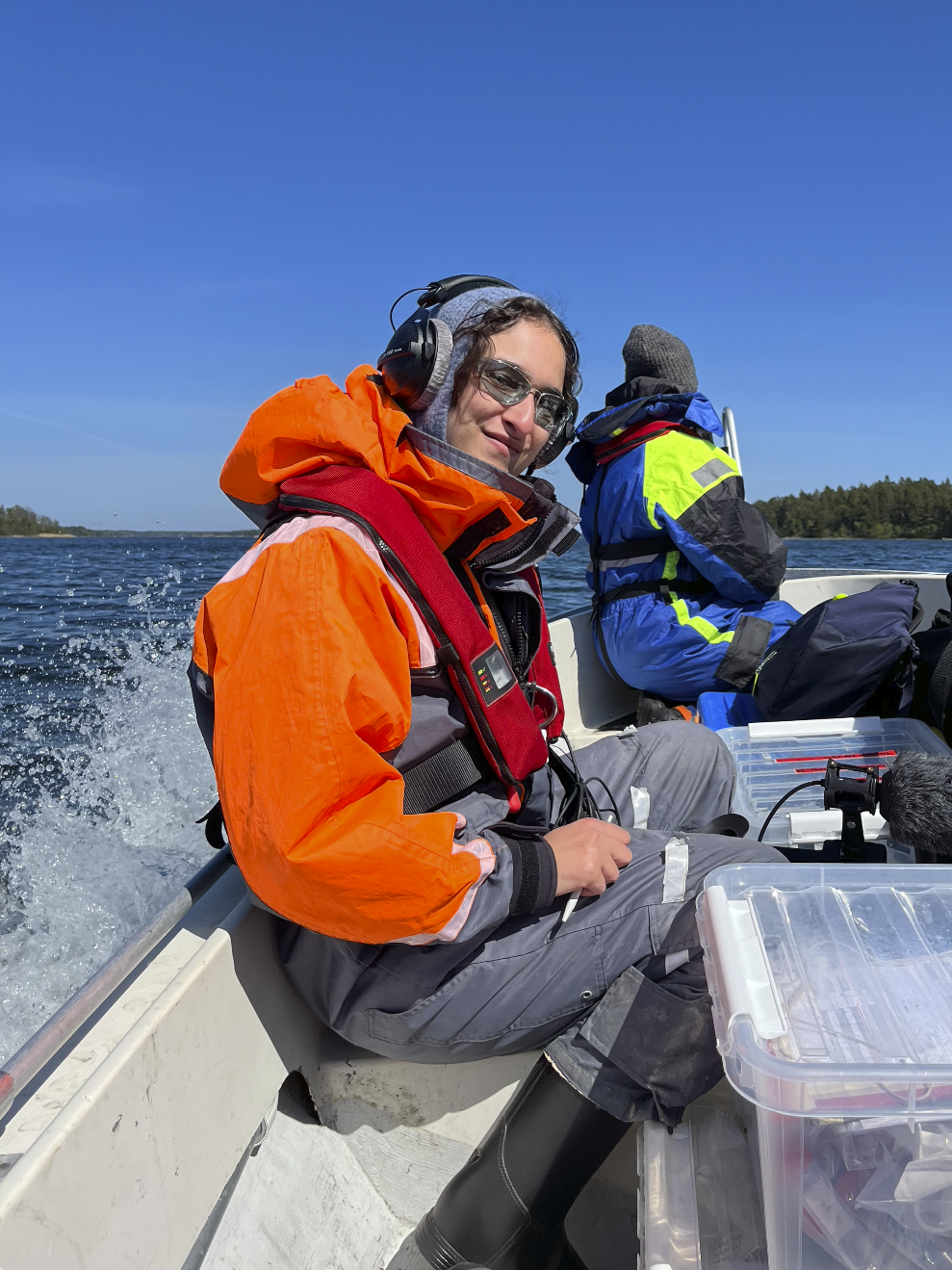 A picture of a person on a small boat in the Archipelago, wearing technical gear and a set of headphones to record sound for a film.