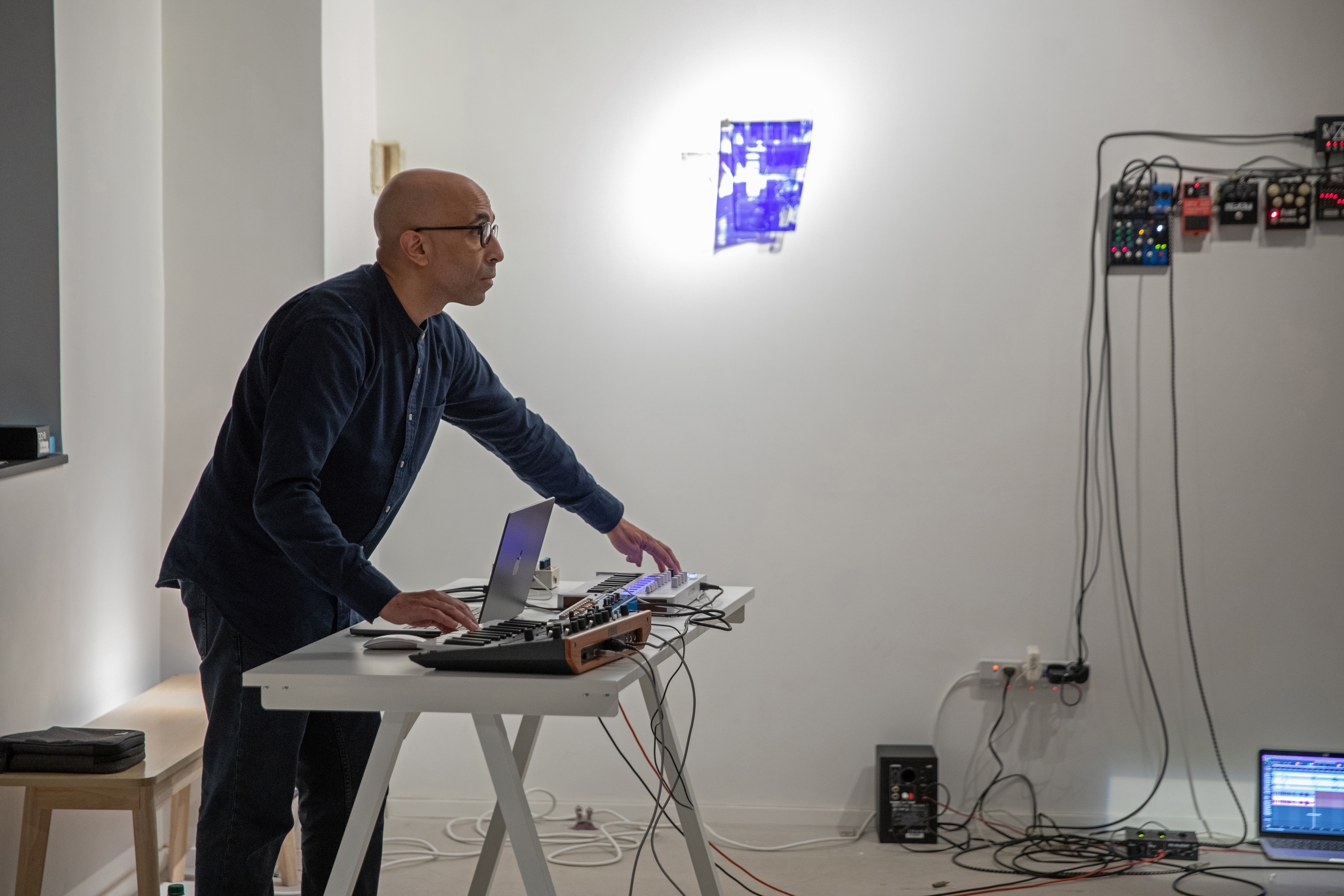 A man with glasses stands at a table using a laptop, keyboard controller, and electronic sound equipment in front of a white wall with cables and mounted devices.