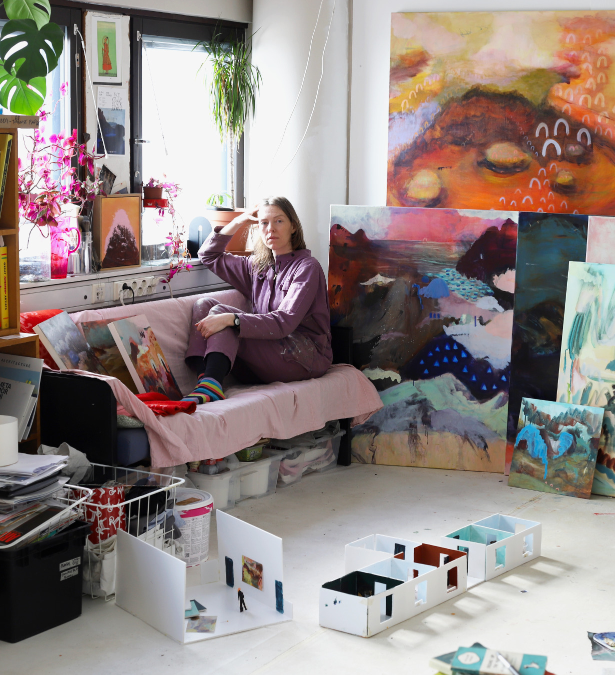 A woman sits on a pale pink-covered sofa in a bright artist’s studio, surrounded by large, colorful abstract landscape paintings leaning against the walls. She wears a mauve paint-splattered outfit and striped socks, resting her head on her hand. Sunlight streams through a window filled with plants. On the floor are small white architectural models and art materials.