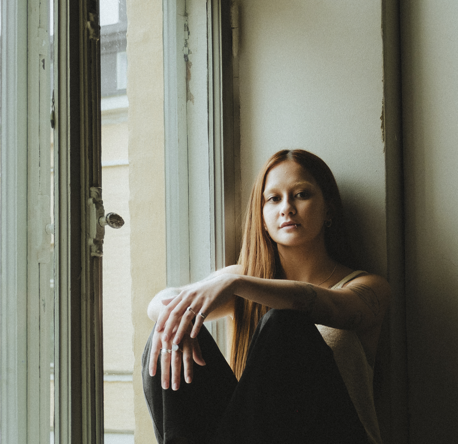 A person with long brown hair is sitting by a window. She is wearing dark pants and a light-colored top and has her hands resting on her knees.